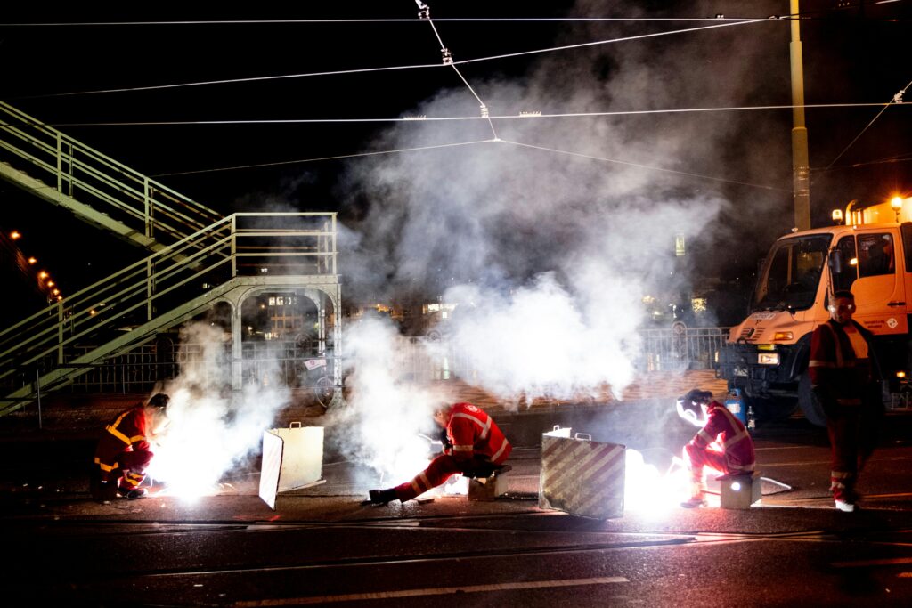 Welders in uniform working on a city street at night amidst smoke and lights.